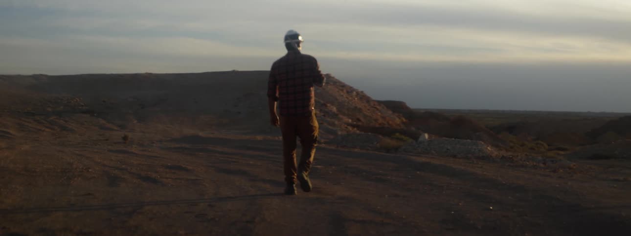 A man wearing a hardhat is walking with a pickax over his shoulder near a dirt mound