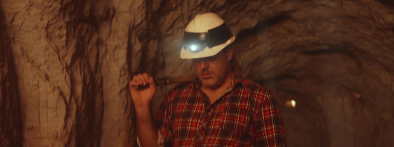A man is walking in an underground mine with a headlamp on his hardhat and holding a pickax on his shoulder