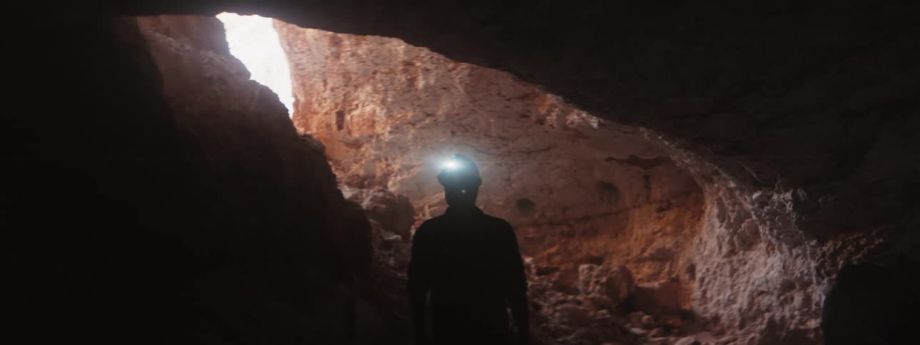 The silhouette of a man standing at the opening of a cavern wearing a helmet with headlamp