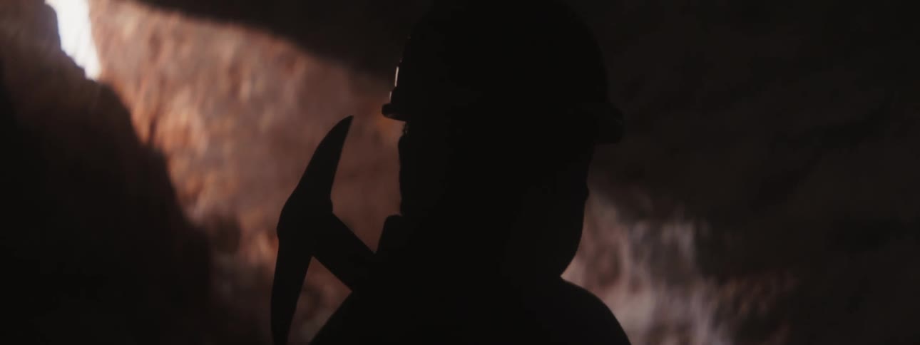 Miner carrying pickax is silhouetted against light entering from mouth of mine