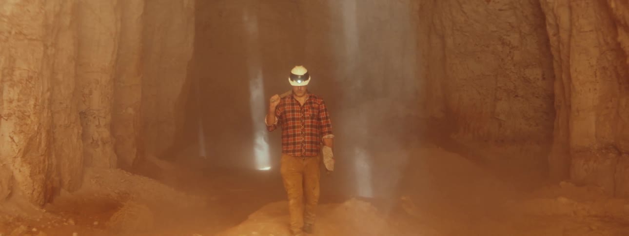 Miner walks through mine carrying shovel and wearing hardhat with headlamp