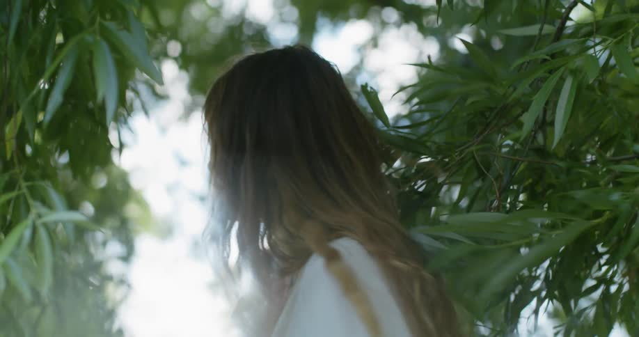 Woman with brown wavy hair and white dress walks among trees