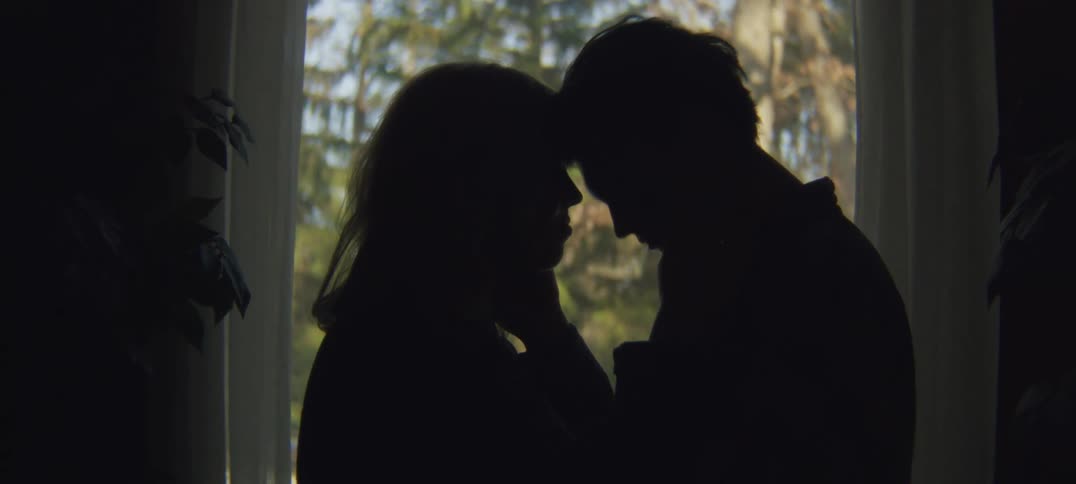 Woman and grieving man holding each other are silhouetted against the window light