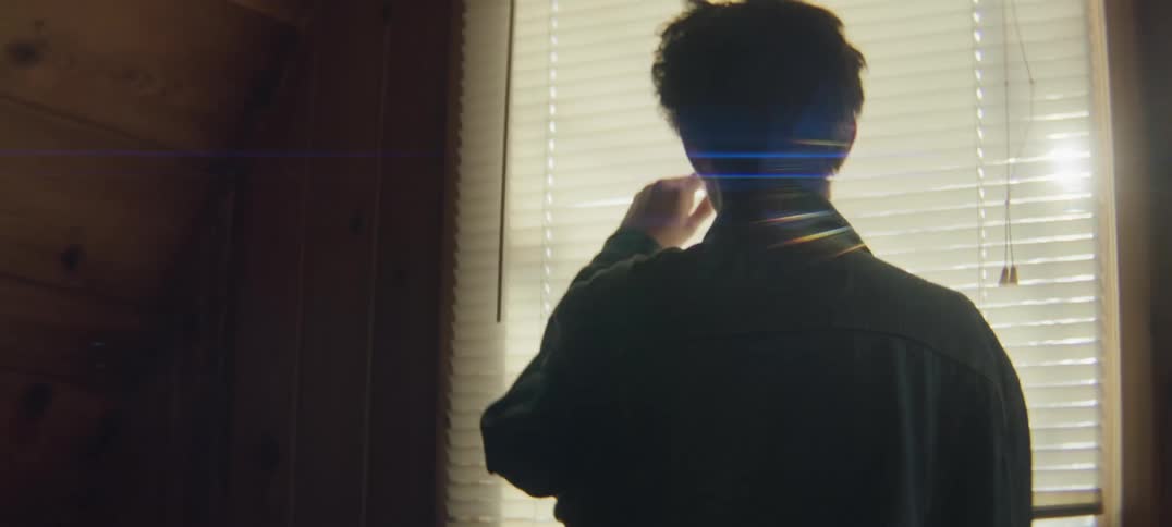 A teenage boy at home turns in circles with his hands behind his head and then peaks out through the blinds on the window