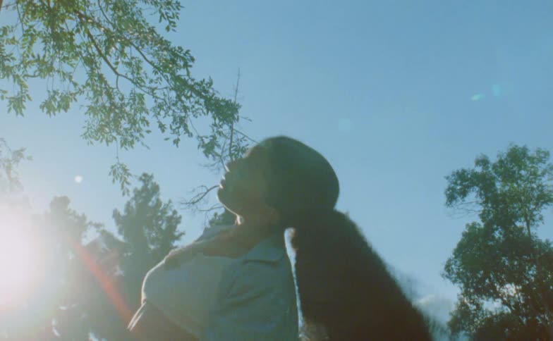 Woman with ponytail blue shirt and white top dances under trees against the blue sky with shining sun