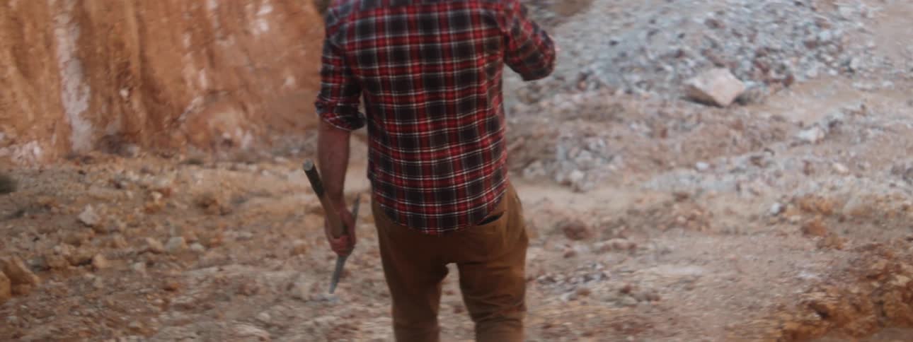Man in plaid shirt and brown pants walks downhill carrying a pickax
