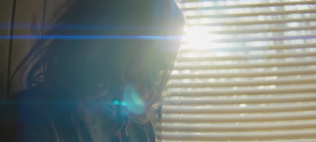 Blonde woman turns her head to look toward blinds that are letting sun glare into room