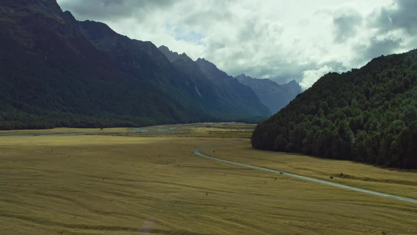 An aerial view of a car driving on a road past a grassland and mountain range