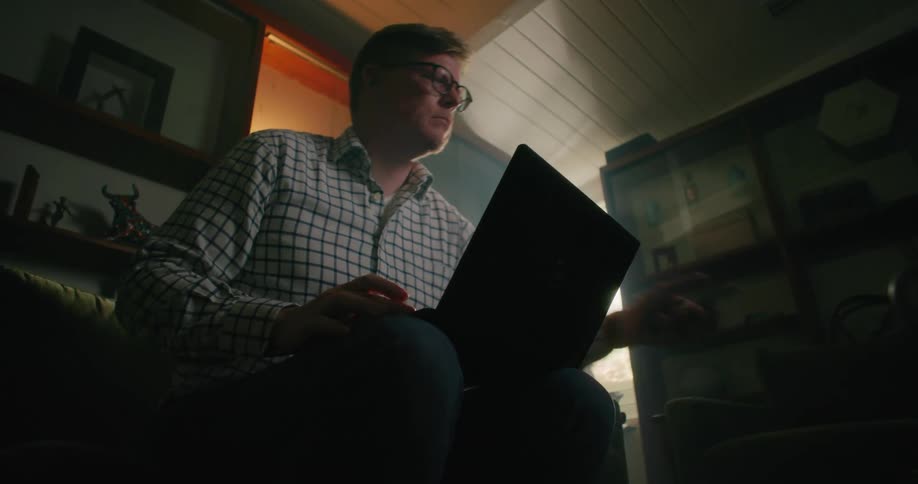 A man is sitting inside a room typing on a laptop computer and he reaches for his cell phone