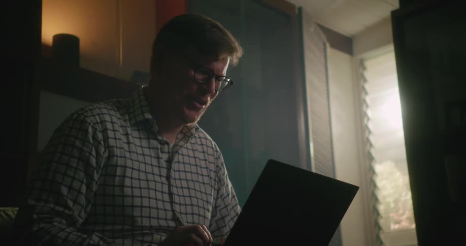A man is sitting inside a room laughing as he types on a laptop computer as sunlight shines in the window