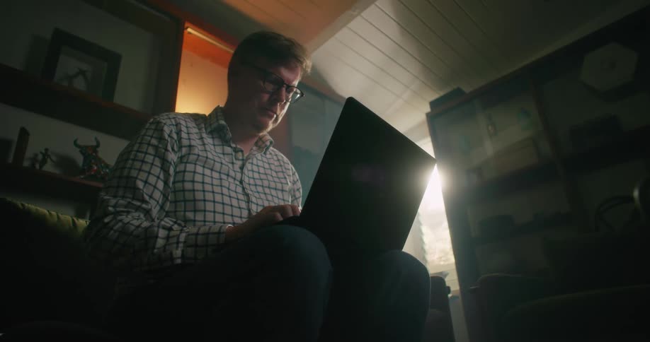 A man is sitting inside a room typing on a laptop computer as sunlight shines in the window