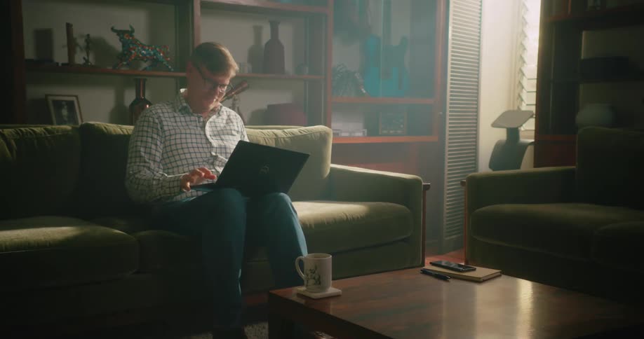 A man sits on a couch at home with a laptop on his knees