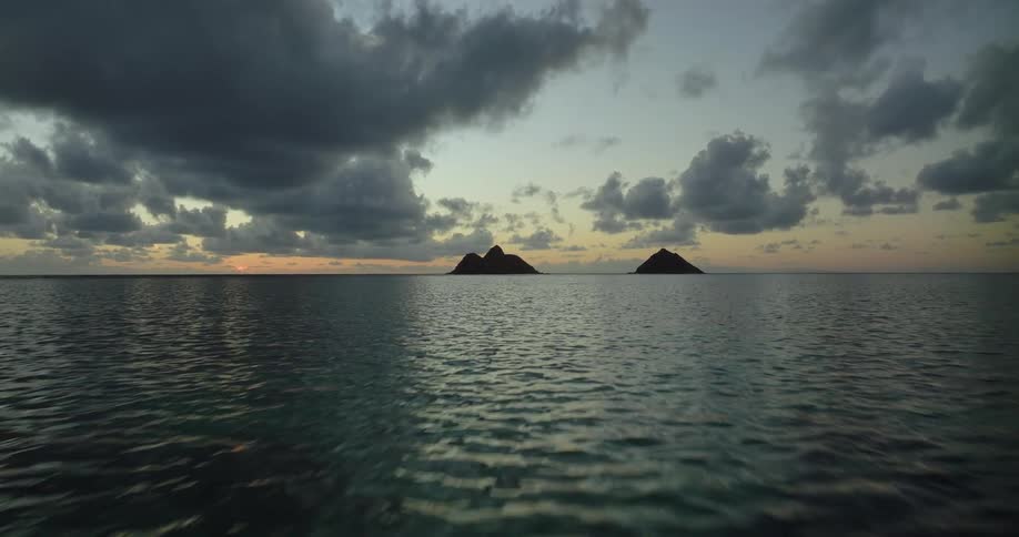 Person wades into ocean across from pair of islands silhouetted against the red horizon on a cloudy day