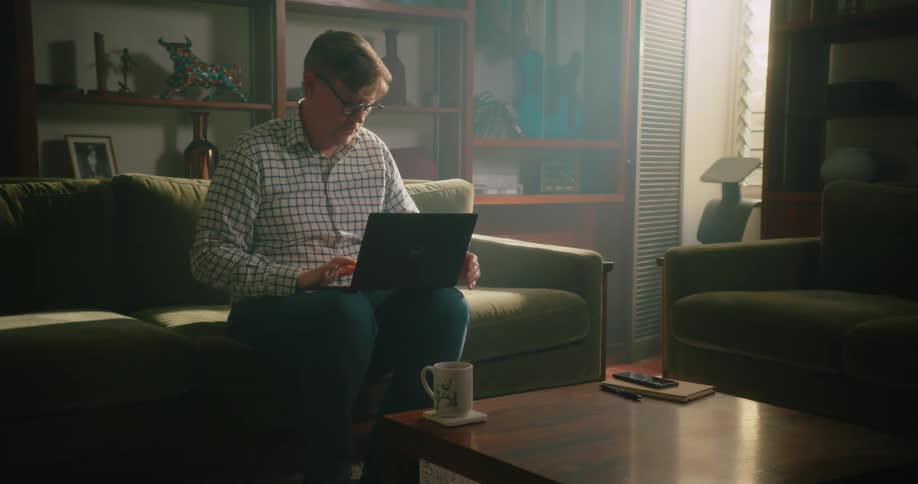 A man is sitting on the couch with his laptop computer on his knees and he opens the lid and starts typing
