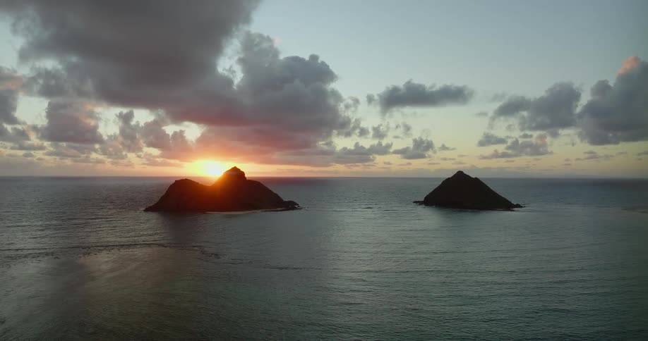 Sunlight shines on the water as it ripples and waves as islands of rock formations are in the distance under a cloudy sky with the sun on the horizon
