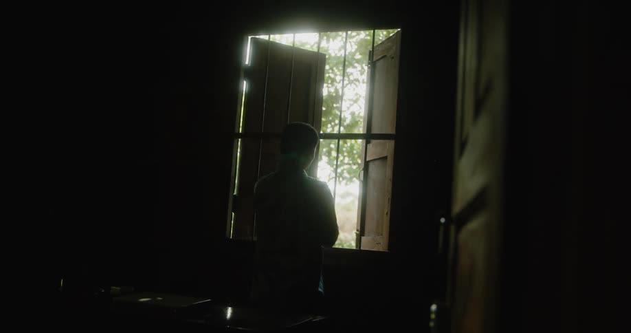 Man opens shutters on barred window letting light brighten dark room