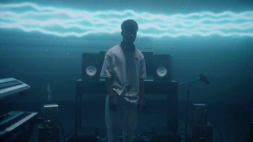 A man with headphones around his neck standing on a small stage surrounded by electronic music equipment and a digital screen