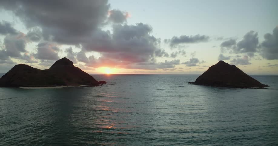 Sunlight shines on the water as it ripples and waves as islands of rock formations are in the distance under a cloudy sky with the sun on the horizon