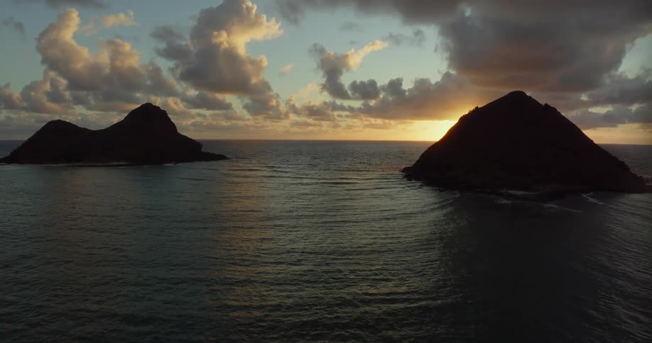 Sunlight shines on the water as it ripples and waves as islands of rock formations are in the distance under a cloudy sky with the sun on the horizon
