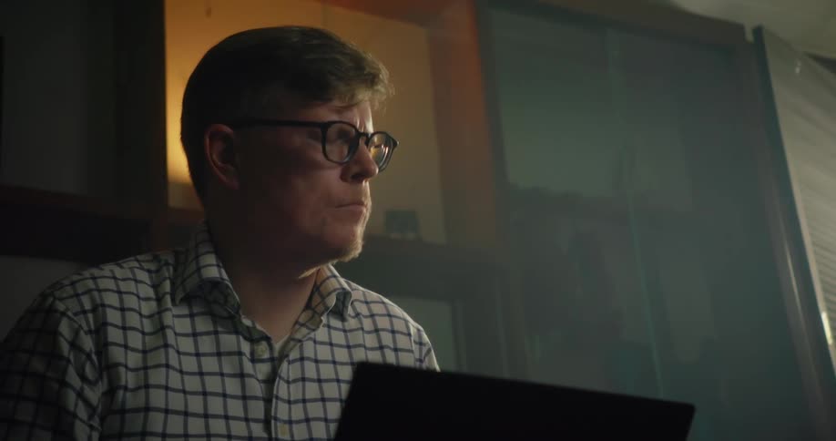 A man wearing glasses is sitting in front of a laptop computer looking up and then he looks back down and types
