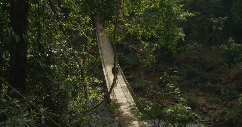 A man walks across a swinging footbridge over a stream in the woods and it moves as he walks