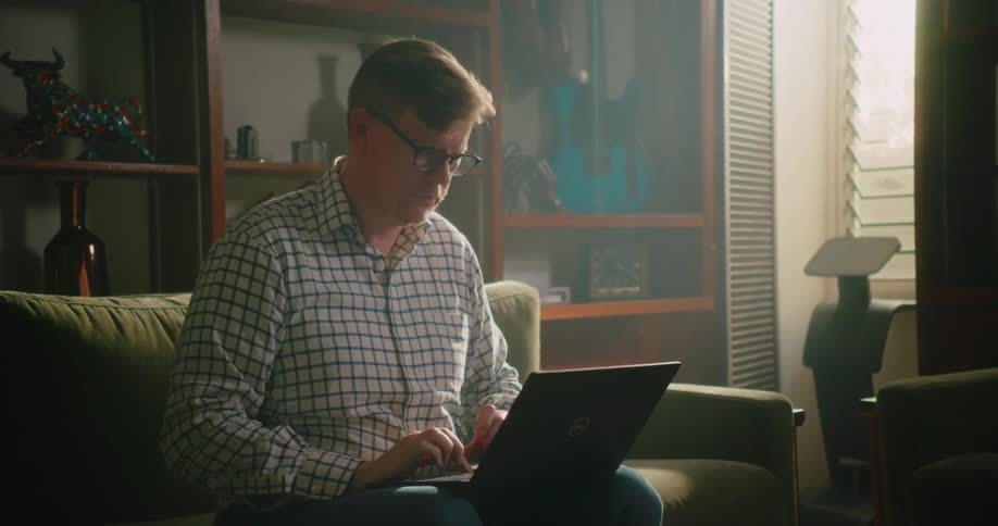 A man wearing glasses is sitting on the couch typing on a laptop computer