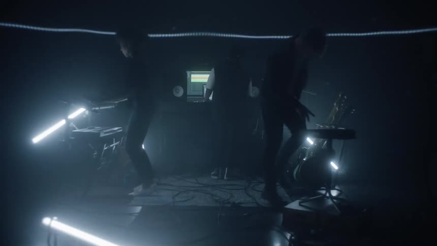 Three people are dancing on a small platform in the dark with flashing lights and musical instruments all around