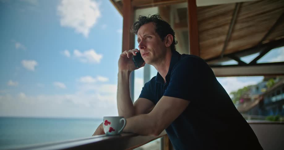 A man talks on the phone while standing on a balcony overlooking the coast