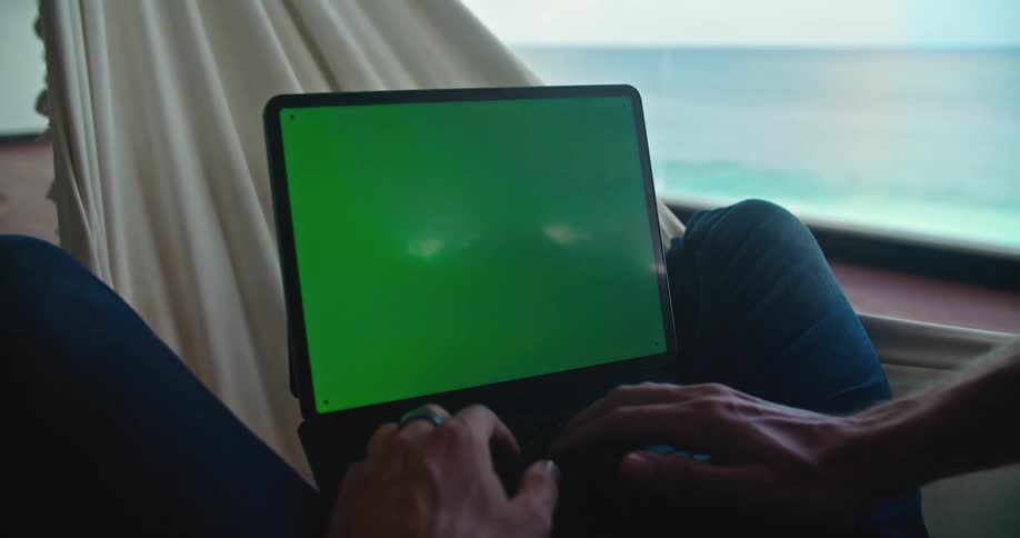A person is sitting on a hammock typing on a computer keyboard and the monitor has a green screen