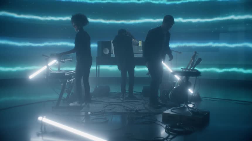 A man and woman walk on to a stage to join another musician in front of a large screen with wavy lines moving across it