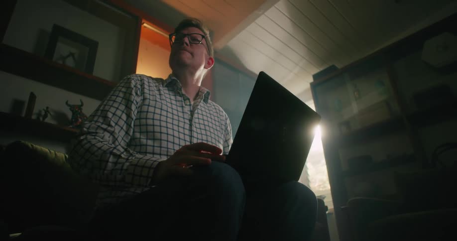 A man is sitting inside a room typing on a laptop computer and he pauses to think as sunlight shines in the window
