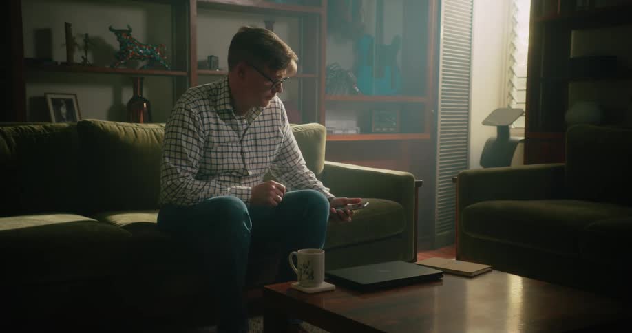 A man walks into the living room with a cup of coffee and he sits down on the couch and reaches into his back pocket and puts his cell phone on the table in front of him next to his laptop computer