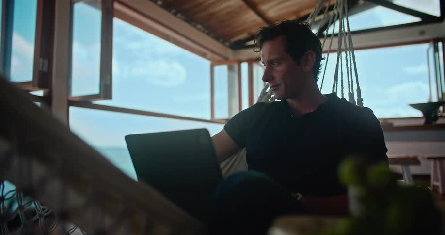 A man sitting on a hammock in a cabin leans back then looks at his laptop with a smile