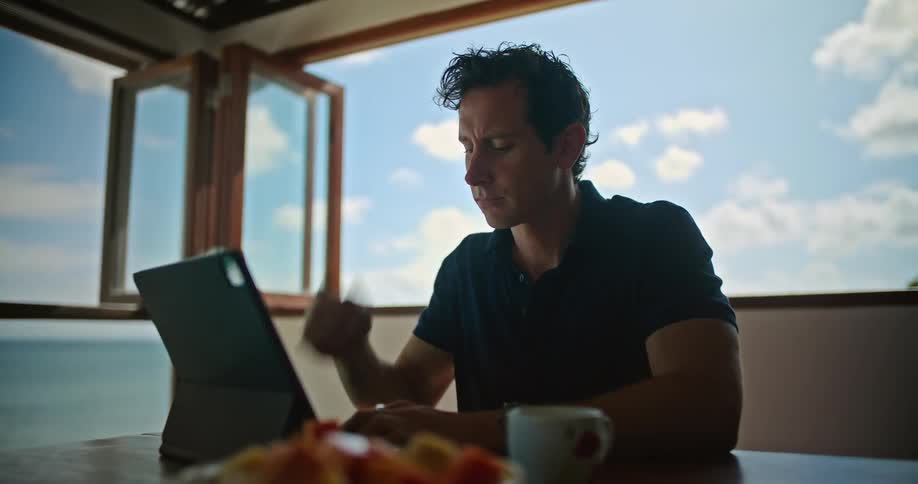 A man is sitting at a table typing on a laptop computer and he stops to drink his coffee