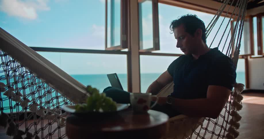 A man is relaxing on the patio on a hammock with his laptop computer and a cup of coffee