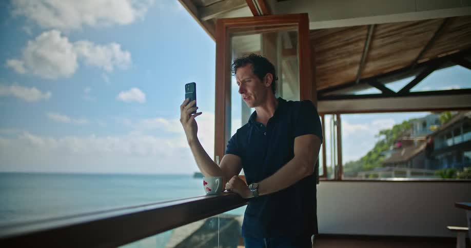 A man drinking coffee stands on his balcony on the shore and video chats with someone on his cell phone as he looks out at the ocean