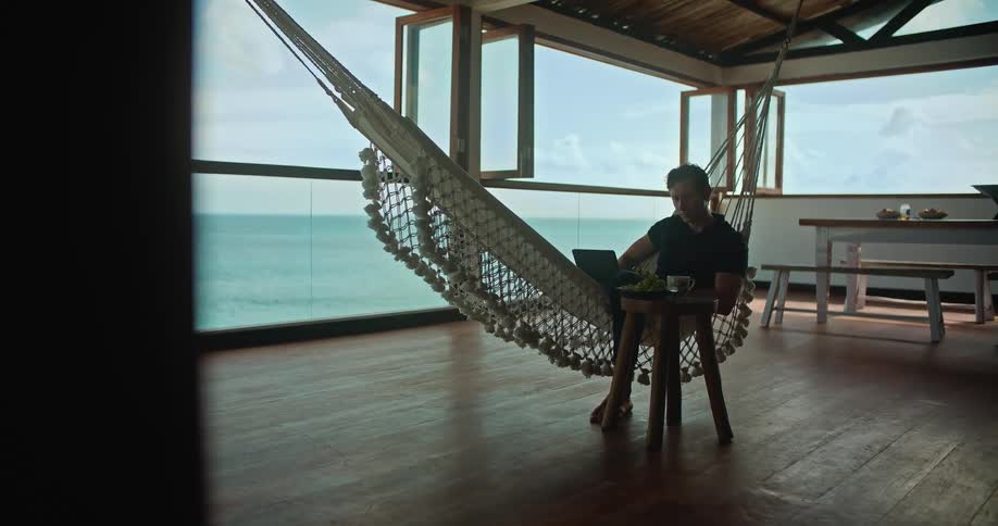 A man works on his laptop while sitting in a hammock on his balcony
