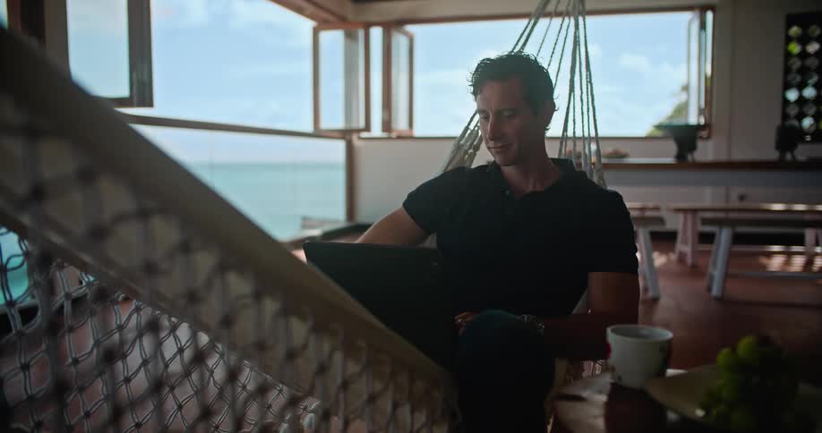 A man sits in his home on a hammock with several windows open to see the beach while on his laptop