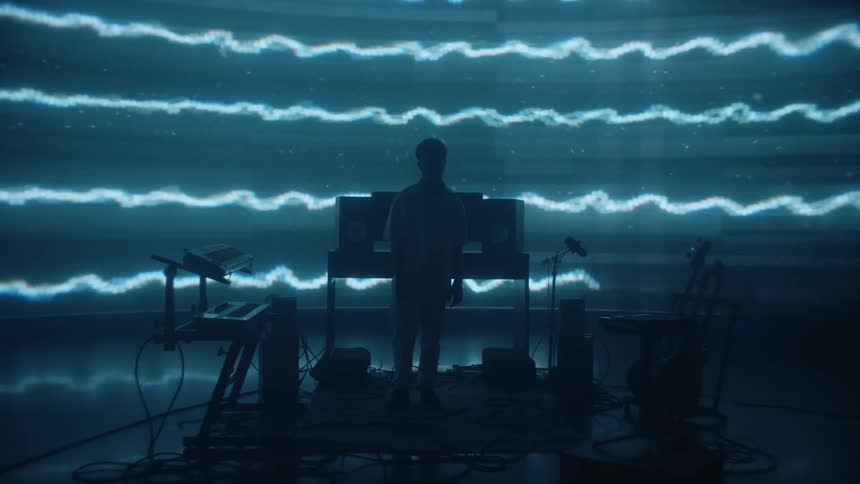 Blue waving lines move across the wall and a silhouette of a man standing in front of computers with keyboards and guitars to each side of him