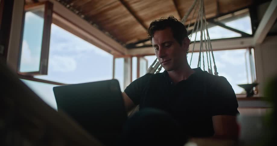 A man is relaxing on the porch on a hammock with his laptop computer and looking out the window with his chin resting on his hand