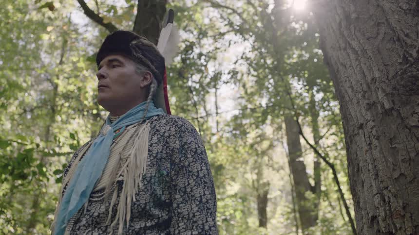 A man with a feather on his hat stands under a tree