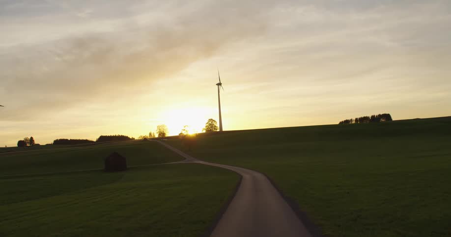 On a rural road the sun shines brightly upon wind turbines spinning on a wind farm
