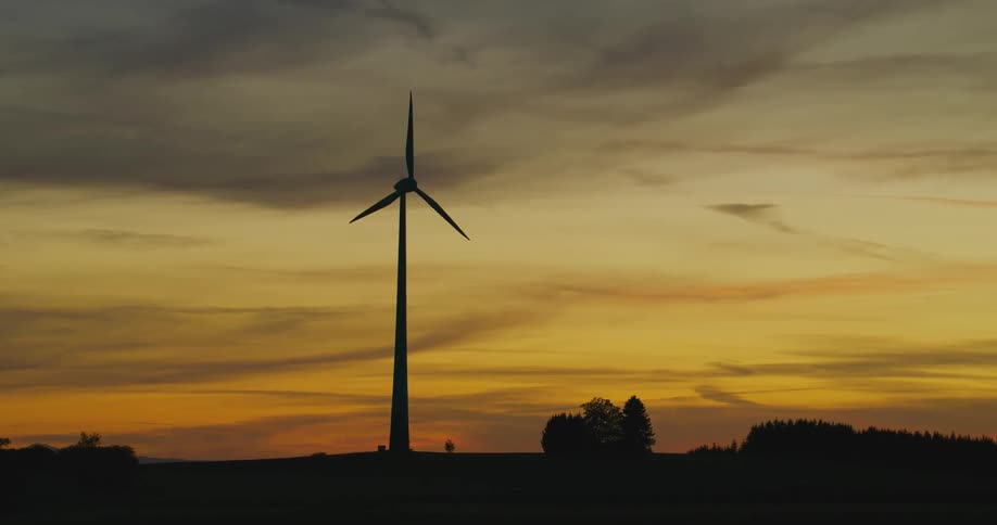 The sun sets in an orange sky as a wind turbine spins high above trees