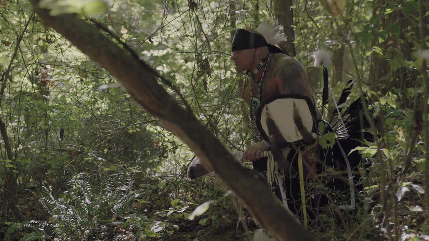 A man wearing a feathered hat looks around the woods