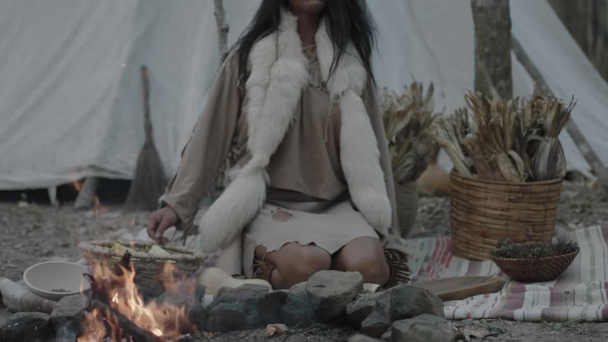 A woman wearing native clothing approaches and sits at a small campfire and starts to prepare food
