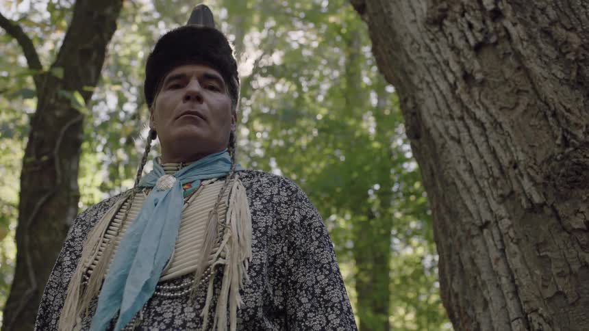 A native american man standing next to a tree looks down towards the ground