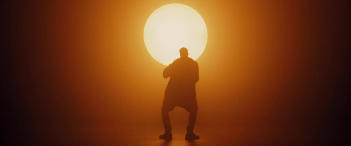 A man wearing a long coat is standing in front a bright sunlike light raises his arms and dances as his silhouette moves like a shadow in the light