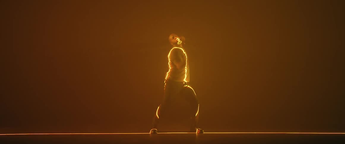 A man is standing in an orange haze and doing martial arts moves as a dance with a yellow light shining on him like lightning