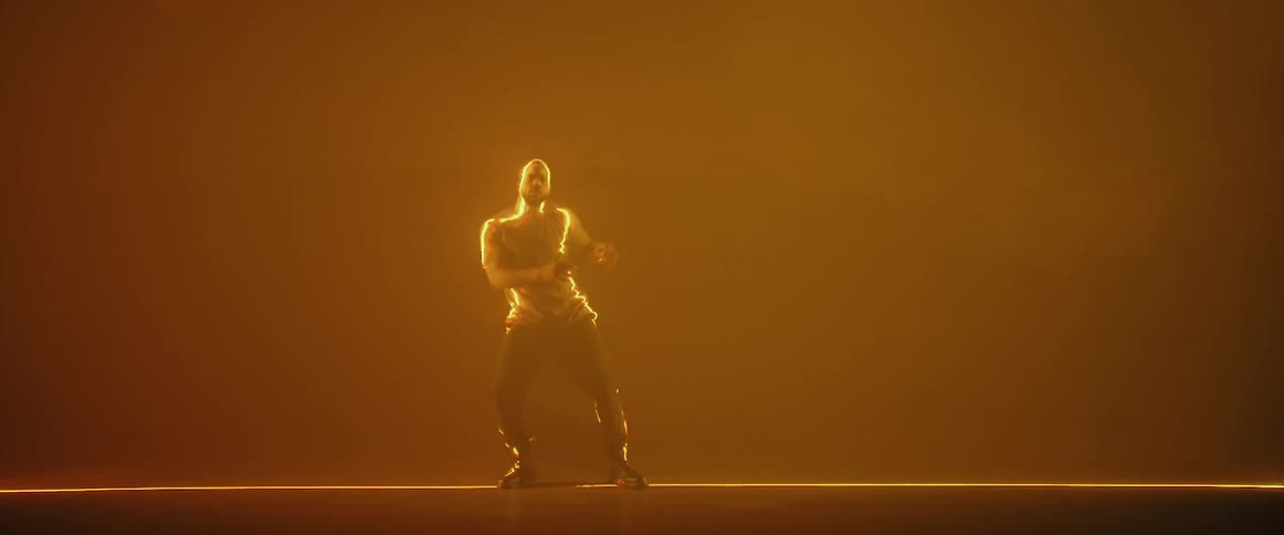 A man is standing on a stage in an orange haze dancing with a yellow light shining on him as he raises his arms in the air