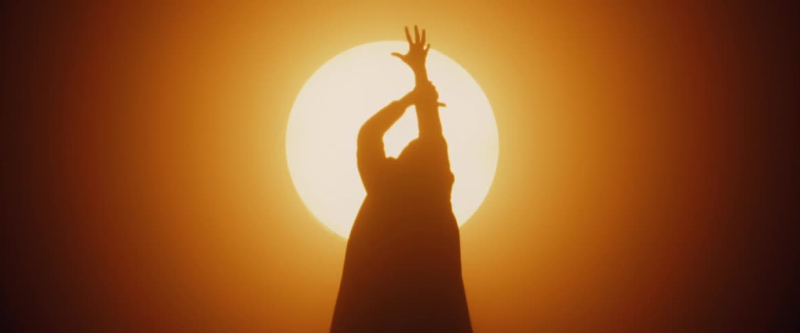 A man wearing a long coat is standing in front a bright sunlike light dancing as his silhouette moves like a shadow in the light